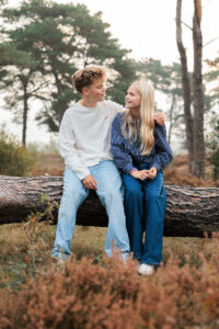 Jesse en Isa zitten samen op een boomstam in het bos en kijken elkaar lachend aan tijdens hun familiefotoshoot. De warme kleuren van de heide en bomen zorgen voor een rustige, natuurlijke sfeer.