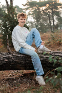 Jesse zit ontspannen op een boomstam en kijkt lachend richting de camera. Een natuurlijk portret uit de familiefotoshoot in de omgeving van Bakkeveen.