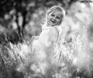 Een zwart-wit foto van een jong meisje met blond haar, gekleed in een wit jurkje, lachend en omringd door hoge grassen en bloemen bij zonsondergang. De zachte belichting en onscherpe achtergrond creëren een dromerige, sprookjesachtige sfeer.