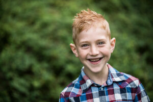 Lachende jongen met kort rood haar en een geruite blouse, staande voor een groene, wazige achtergrond.