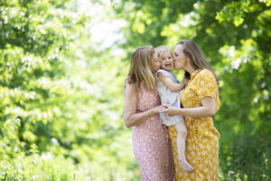 Drie generaties vrouwen in een groene omgeving: een grootmoeder en moeder kussen een jong meisje, allemaal gekleed in kleurrijke jurken met bloemenpatroon, met zonlicht dat door de bomen schijnt.