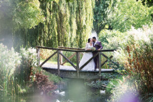 Bruid en bruidegom delen een kus op een houten brug omgeven door weelderige groene vegetatie en een serene vijver, met zonlicht dat door de bladeren schijnt.