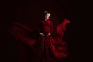 Zwanger vrouw poseert in een dramatische rode jurk met vloeiende stof die om haar heen waait, tegen een donkere achtergrond. Ze houdt haar handen liefdevol op haar buik en kijkt neer met een serene glimlach.