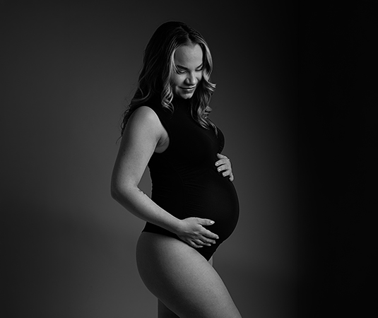 Zwangere vrouw staat in een zwarte body in een fotostudio ze heeft haar handen op haar buik en lacht naar haar zwangere buik.