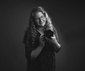 Fotografe Christha met bril en krullen houdt camera vast tijdens portretshoot in zwart-wit