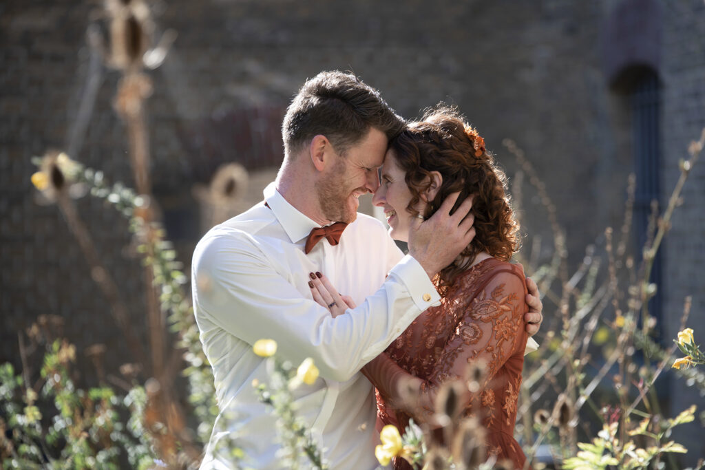 Bruid en bruidegom lachen en drukken hun voorhoofden tegen elkaar, een speels en intiem moment in de tuin.