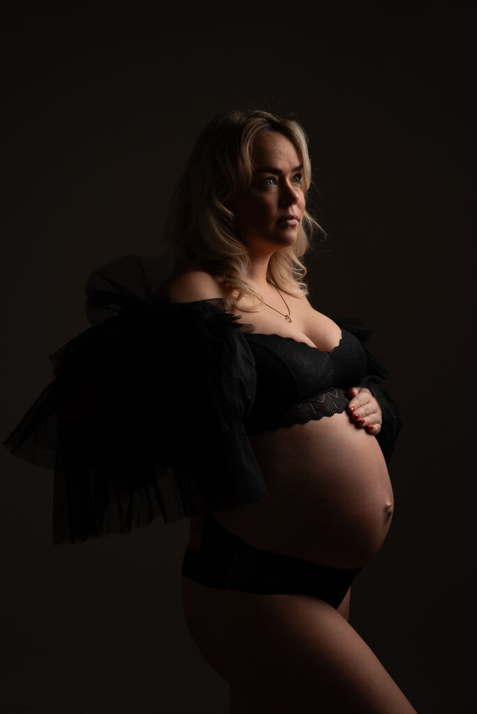 Zwangere vrouw in zwarte lingerie met transparante mouwen tijdens een zwangerschapsshoot in een fotostudio in Leeuwarden met donker contrastlicht.