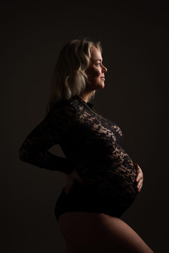 Zijaanzicht van een zwangere vrouw in zwarte kanten body tijdens een studio zwangerschapsshoot in Leeuwarden met zacht schaduwlicht.