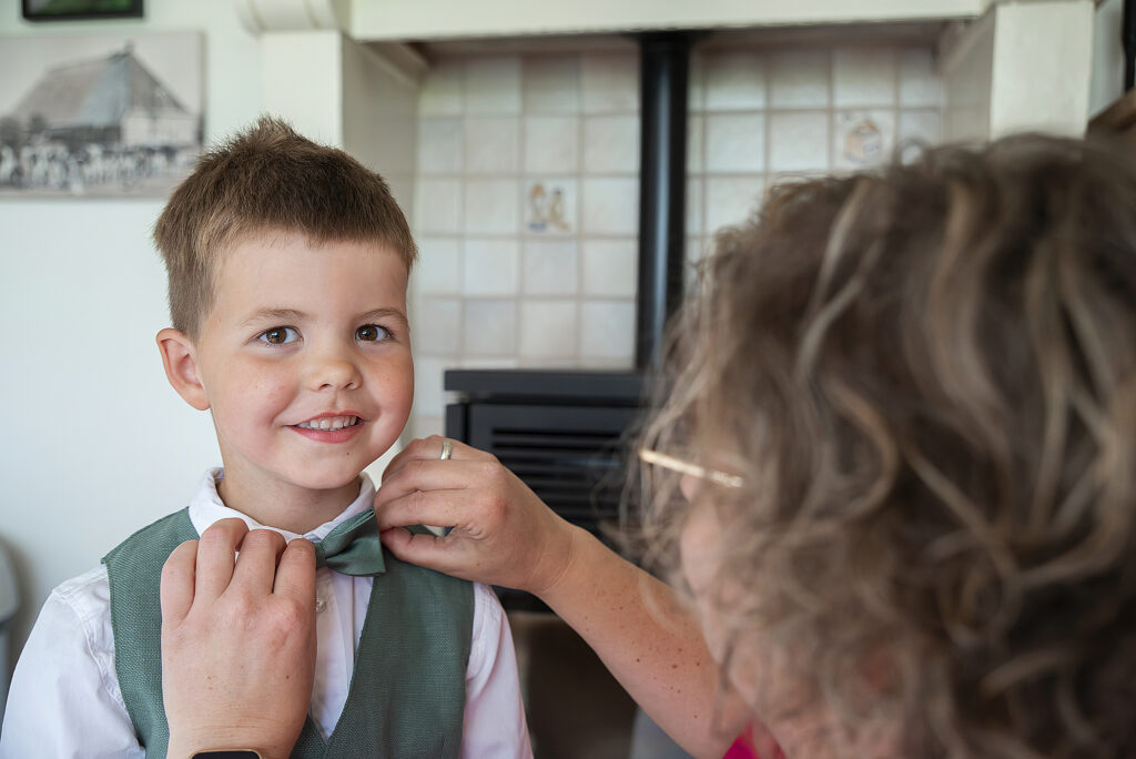 Een jonge jongen met een brede glimlach draagt een witte blouse met een groene gilet en een vlinderdas, terwijl hij wordt klaargemaakt voor een bruiloft. Een volwassene past zijn vlinderdas aan. Op de achtergrond is een betegelde muur en een deel van een haard zichtbaar.