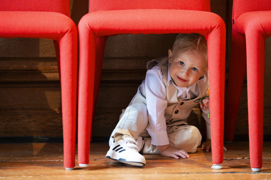 Een jong kind, gekleed in een nette outfit met witte Adidas-schoenen, kijkt glimlachend onder een rij felrode stoelen door.