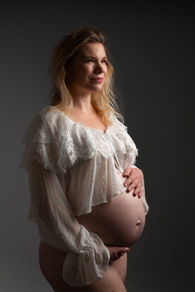 Zwangere vrouw straalt in witte kanten blouse tijdens zwangerschaps shoot bij Fotografe Christha.