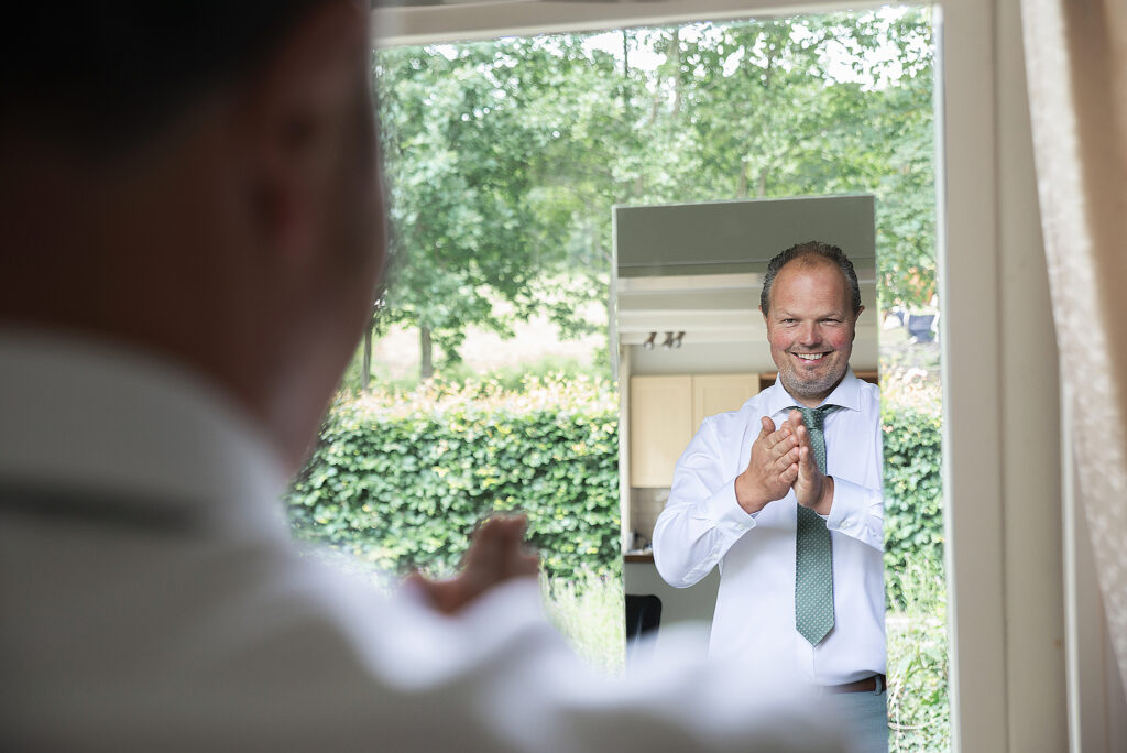 Een man, gekleed in een witte blouse met een groene stropdas, staat voor een spiegel en klapt in zijn handen terwijl hij zich voorbereidt op zijn trouwdag. Hij kijkt glimlachend naar zijn reflectie. De achtergrond toont een buitenruimte met groene bomen en struiken. De man wordt deels vanuit een hoek gefotografeerd, waarbij zijn reflectie centraal in beeld is.