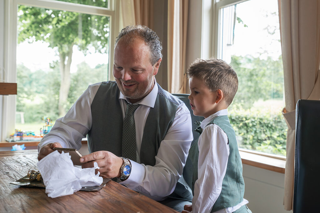 Een man en een jongen, beiden gekleed in groene gilets en witte overhemden, zitten samen aan een tafel en openen een cadeau. De man glimlacht terwijl hij het cadeau uitpakt, terwijl de jongen geïnteresseerd toekijkt. Op de achtergrond zijn grote ramen met uitzicht op een groene buitenomgeving te zien. De foto is genomen op de ochtend van een trouwdag, waarbij vader en zoon zich samen voorbereiden.