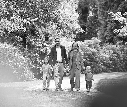 Een zwart-wit foto van een gezin dat samen een wandeling maakt op een bosachtig pad. De ouders lopen hand in hand met hun twee jonge kinderen, omringd door weelderig groen en bomen. De ontspannen en gelukkige gezichtsuitdrukkingen van het gezin stralen warmte en verbondenheid uit.