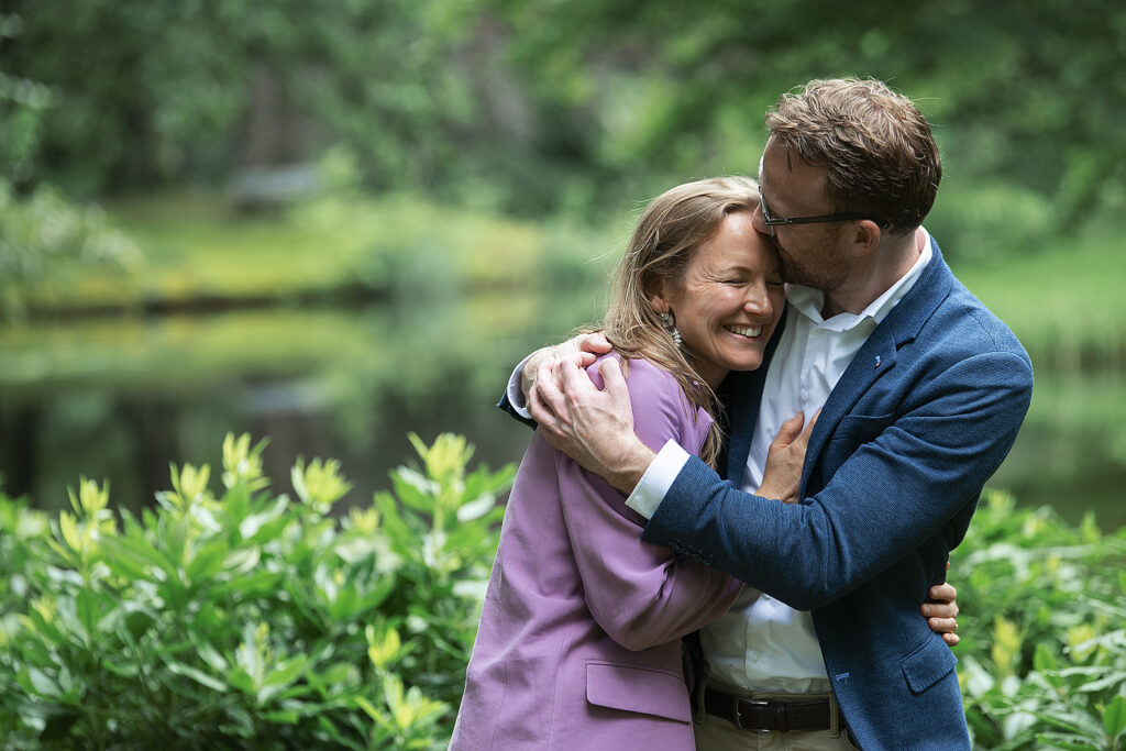 Een romantische foto van een stel dat elkaar omhelst en lacht bij een vijver in het Vijverburgpark, met groene struiken en bomen op de achtergrond.