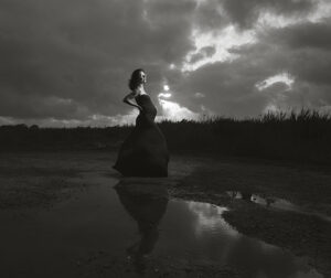 Zwangere vrouw in zwarte jurk poseert krachtig bij dramatische lucht in zwart-wit