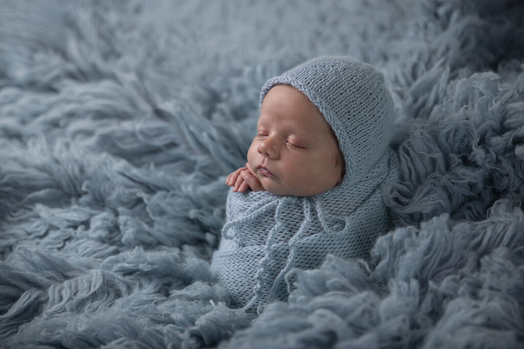 Een foto van een slapende pasgeboren baby, ingewikkeld in een lichtblauwe gebreide deken en muts. De baby ligt op een zachte, pluizige blauwe ondergrond, wat een serene en kalme sfeer creëert.