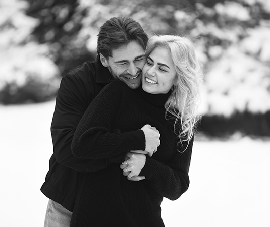 Een zwart-witfoto van een verliefd stel in een besneeuwd landschap. De man omhelst de vrouw van achteren, terwijl ze beiden breed glimlachen. De vrouw heeft lang, golvend blond haar en draagt een zwarte coltrui, terwijl de man een zwarte jas draagt. De achtergrond is zacht vervaagd, waardoor de focus volledig op hun warme en liefdevolle moment ligt.