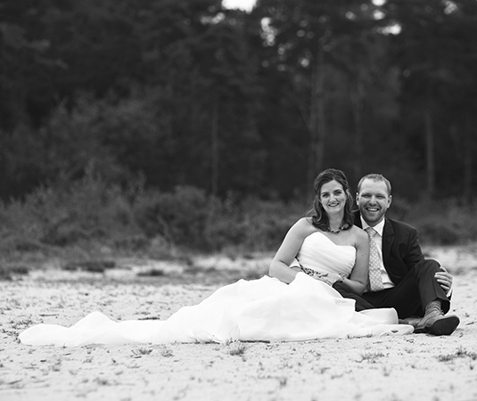 Bruidspaar zit in het zand met bos op de achtergrond, de foto is zwart wit.