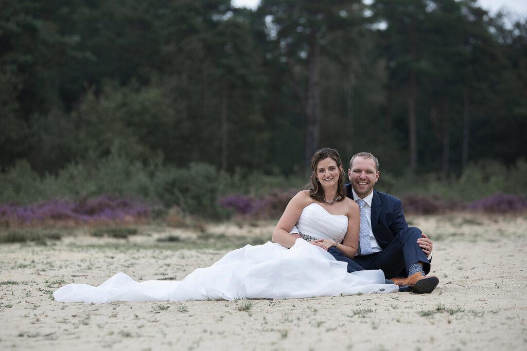 Bruidspaar zit samen in het zand, met in de achtergrond bos en heide.