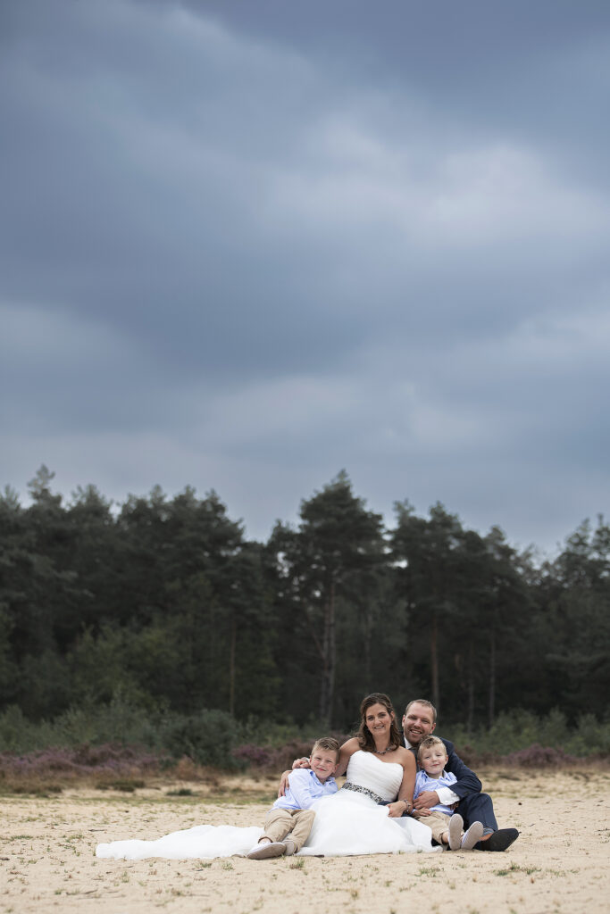 Bruidspaar en hun twee zoons zitten samen in het zand in een natuurgebied met op de achtergrond het bos.