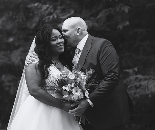 Bruid en bruidegom delen een intiem moment tijdens hun trouwdag. De bruidegom geeft een liefdevolle kus op het voorhoofd van de bruid terwijl ze elkaar innig vasthouden. De bruid houdt een prachtig boeket bloemen vast, en beide stralen van geluk.