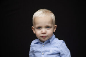 Portret van een jong blond jongetje met een blauwe blouse, kijkend naar de camera tegen een zwarte achtergrond.