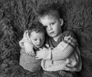 Zwart-witte foto van grote broer Luca die zijn babybroertje Twan omhelst op een zacht wollen kleed, Twan ligt gewrapt in een gebreide sjaal.
