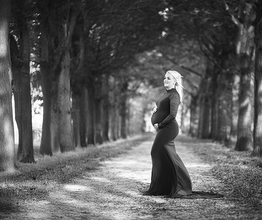Zwangere vrouw in een lange, aansluitende zwangerschapsjurk, poserend op een boslaan met hoge bomen in Leeuwarden, vastgelegd tijdens een zwangerschapsfotoshoot in het zwart-wit.