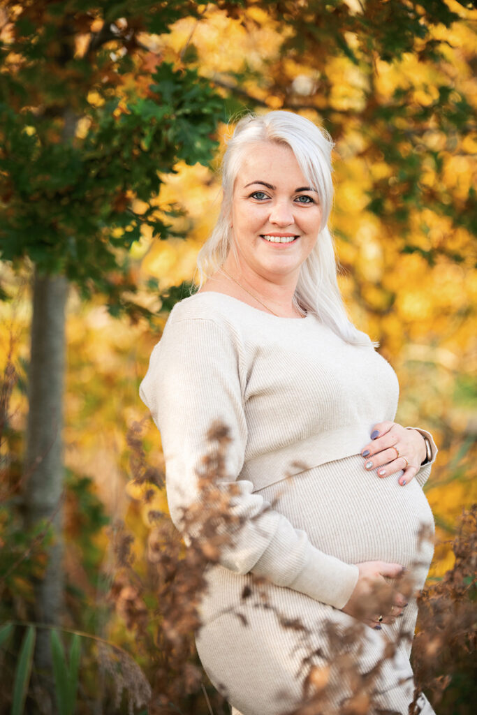 Een warm en puur portret van Michelle in haar eigen comfortabele gebreide set. Ze staat tussen de kleurrijke herfstbladeren, wat een zacht en intiem beeld oplevert. De natuurlijke setting versterkt het rustige en liefdevolle karakter van de foto.