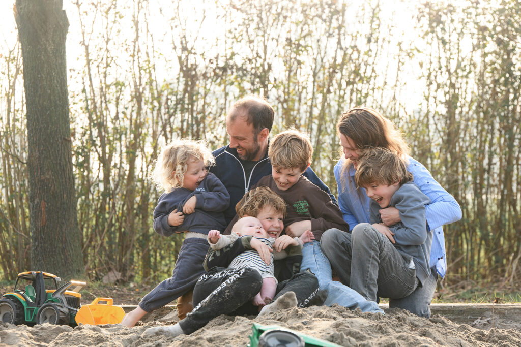 Gezin met vijf kinderen lacht samen in de zandbak tijdens een fotoshoot op hun boerderij