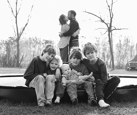 Kinderen van Boer Bart zitten samen op een trampoline terwijl hun ouders elkaar kussen op de achtergrond tijdens een gezinsfotoshoot op de boerderij