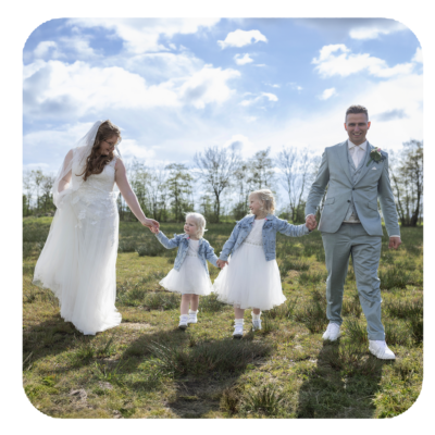 Fotografe Christha met haar dochters en man op hun bruiloft, wandelend hand in hand door een grasveld op een zonnige dag met een blauwe hemel en witte wolken. Christha draagt een witte trouwjurk, de dochters zijn gekleed in witte jurkjes met spijkerjasjes, en haar man draagt een lichtgrijs pak.