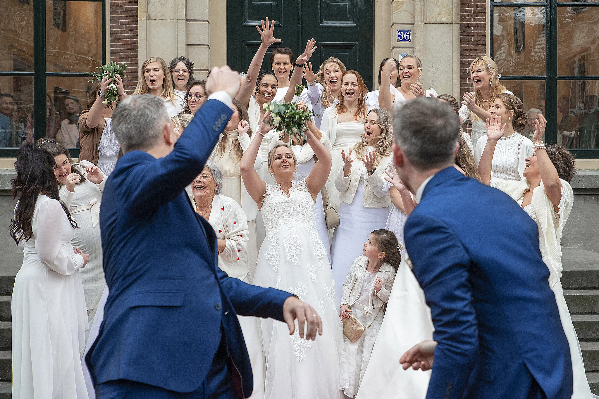 Een vrouw in een witte trouwjurk strekt haar armen uit om een bruidsboeket te vangen, omringd door andere vrouwen die enthousiast toekijken.