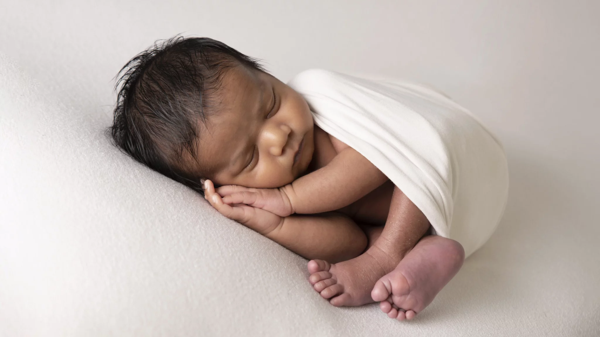 Een pasgeboren baby ligt vredig te slapen, gewikkeld in een witte doek. De baby ligt op zijn zij met zijn handjes onder zijn hoofdje en zijn voetjes ingetrokken, wat een gevoel van geborgenheid en rust uitstraalt. De zachte, lichtgekleurde achtergrond en de delicate belichting benadrukken de tederheid van de slapende baby, wat een serene en pure sfeer creëert.