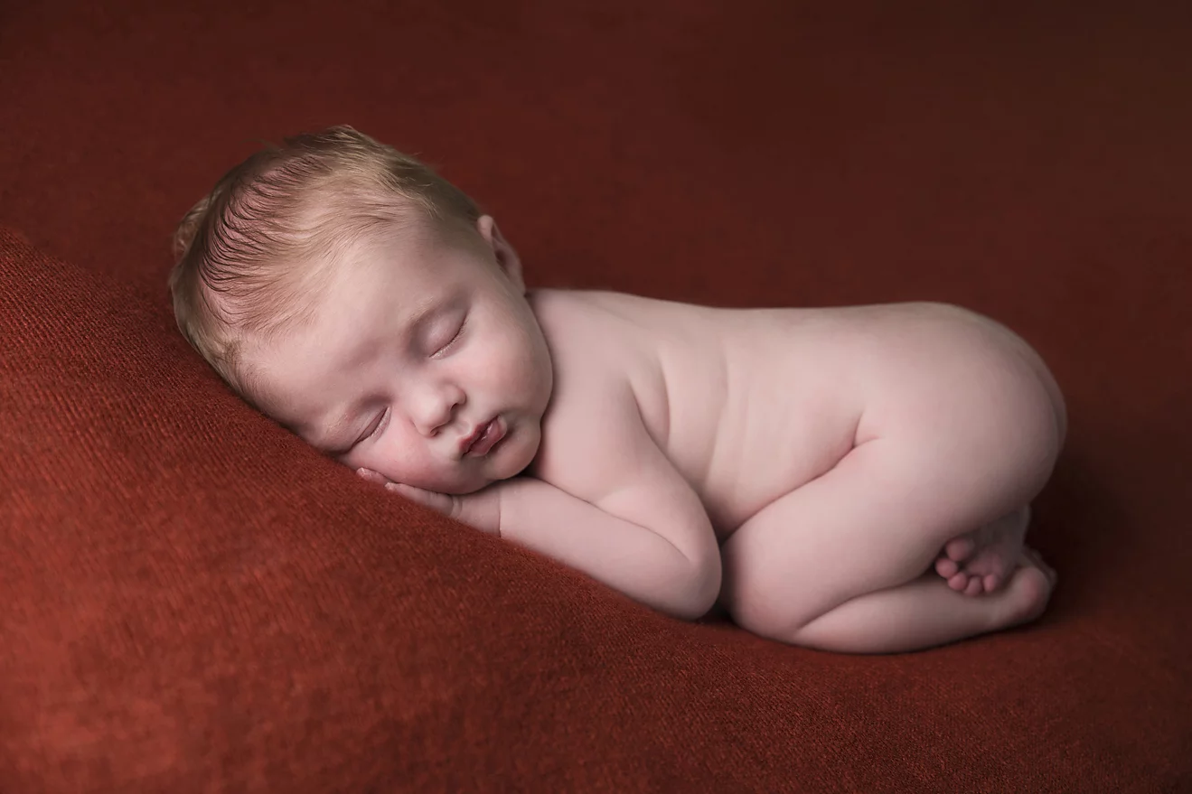 Een slapende pasgeboren baby ligt op zijn zij op een zachte, roestkleurige deken. De baby heeft een rustige uitdrukking op zijn gezicht, met zijn hoofdje rustend op zijn hand en zijn knietjes licht opgetrokken naar zijn buik. De warme kleur van de deken en de zachte belichting zorgen voor een serene en vredige sfeer, waarbij de natuurlijke schoonheid van de slapende baby centraal staat.
