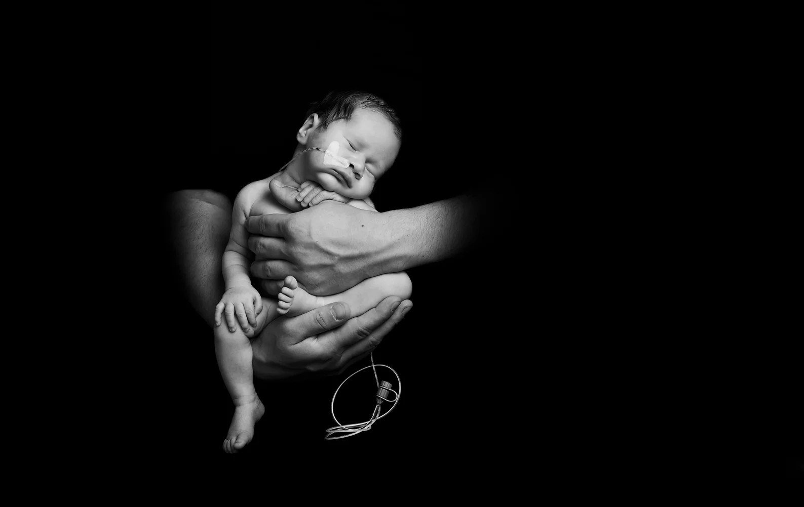 Een ontroerende zwart-wit foto toont een pasgeboren baby die slapend in de handen van een volwassene ligt. De baby heeft een sonde in zijn neus en ligt met zijn hoofdje tegen de borst van de volwassene. De grote handen ondersteunen voorzichtig het kleine lichaam van de baby, wat een gevoel van kwetsbaarheid en bescherming uitstraalt. De donkere achtergrond zorgt ervoor dat de focus volledig op de baby en de handen ligt, wat een serene en emotionele sfeer creëert.