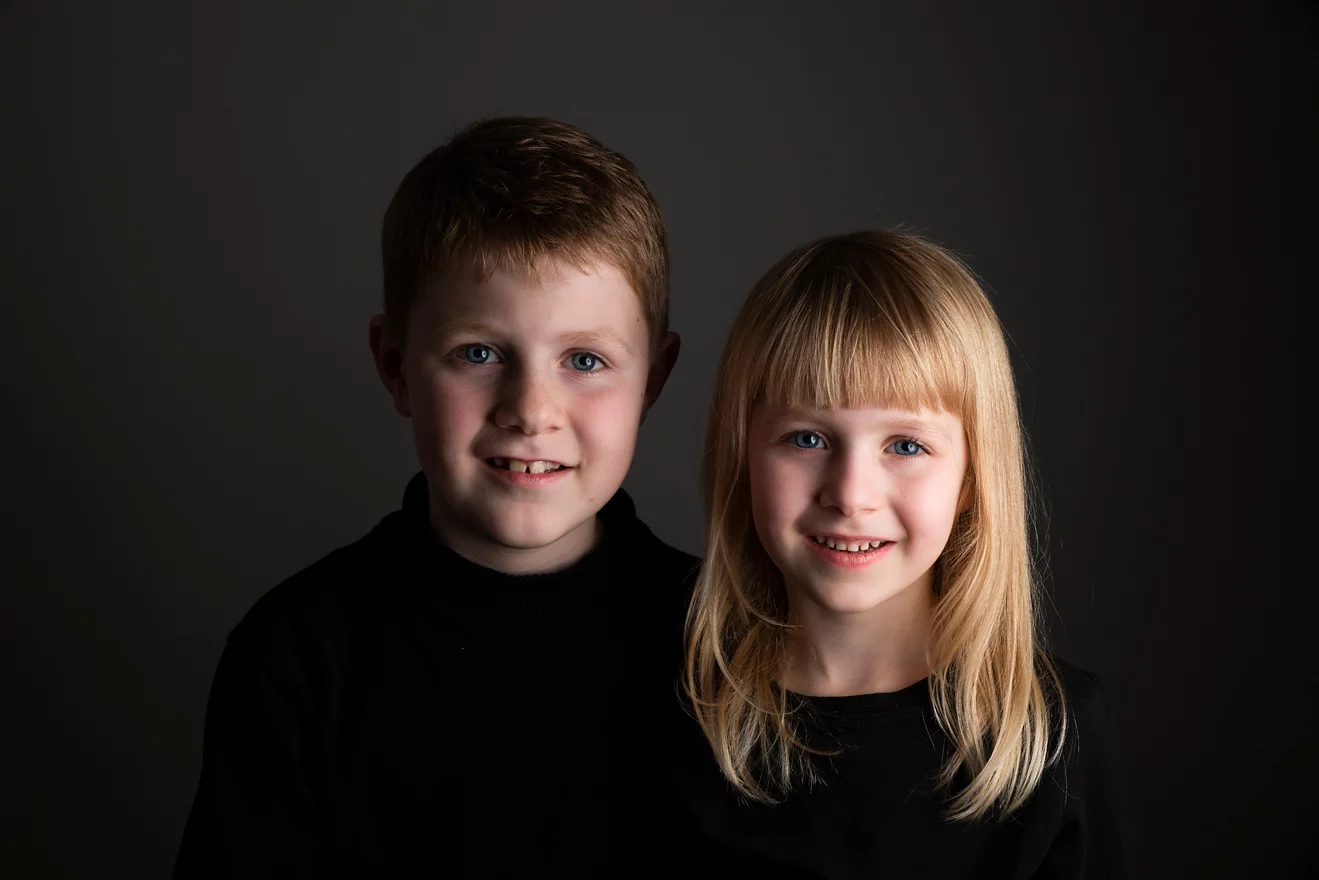 Een portretfoto van twee kinderen voor een donkere achtergrond. Links is een jongen met kort, bruin haar te zien, die een zwarte trui draagt en vriendelijk glimlacht naar de camera. Rechts staat een meisje met blond, halflang haar en een pony, die ook een zwarte trui draagt en breed glimlacht. De belichting benadrukt hun gezichten en zorgt voor een warme, intieme sfeer.