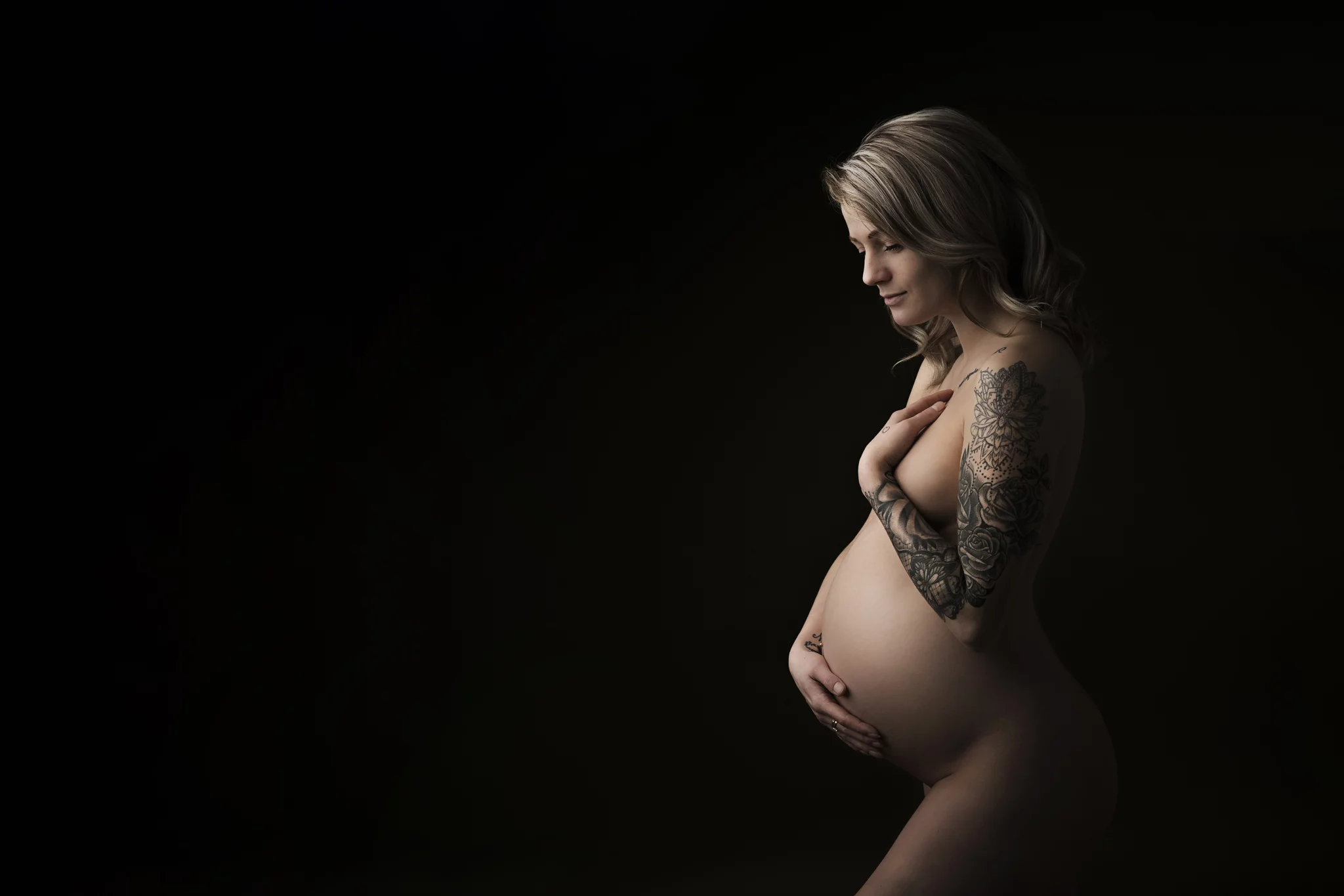 Een zwangere vrouw staat zijwaarts tegen een donkere achtergrond, waarbij haar blote buik prominent zichtbaar is. Ze heeft een grote tatoeage op haar linkerarm die in detail te zien is. Ze houdt één hand beschermend onder haar buik en de andere hand over haar borst. Haar blonde haar valt los over haar schouders en ze kijkt rustig en liefdevol naar beneden. De belichting benadrukt de contouren van haar lichaam, wat een serene en intieme sfeer creëert.
