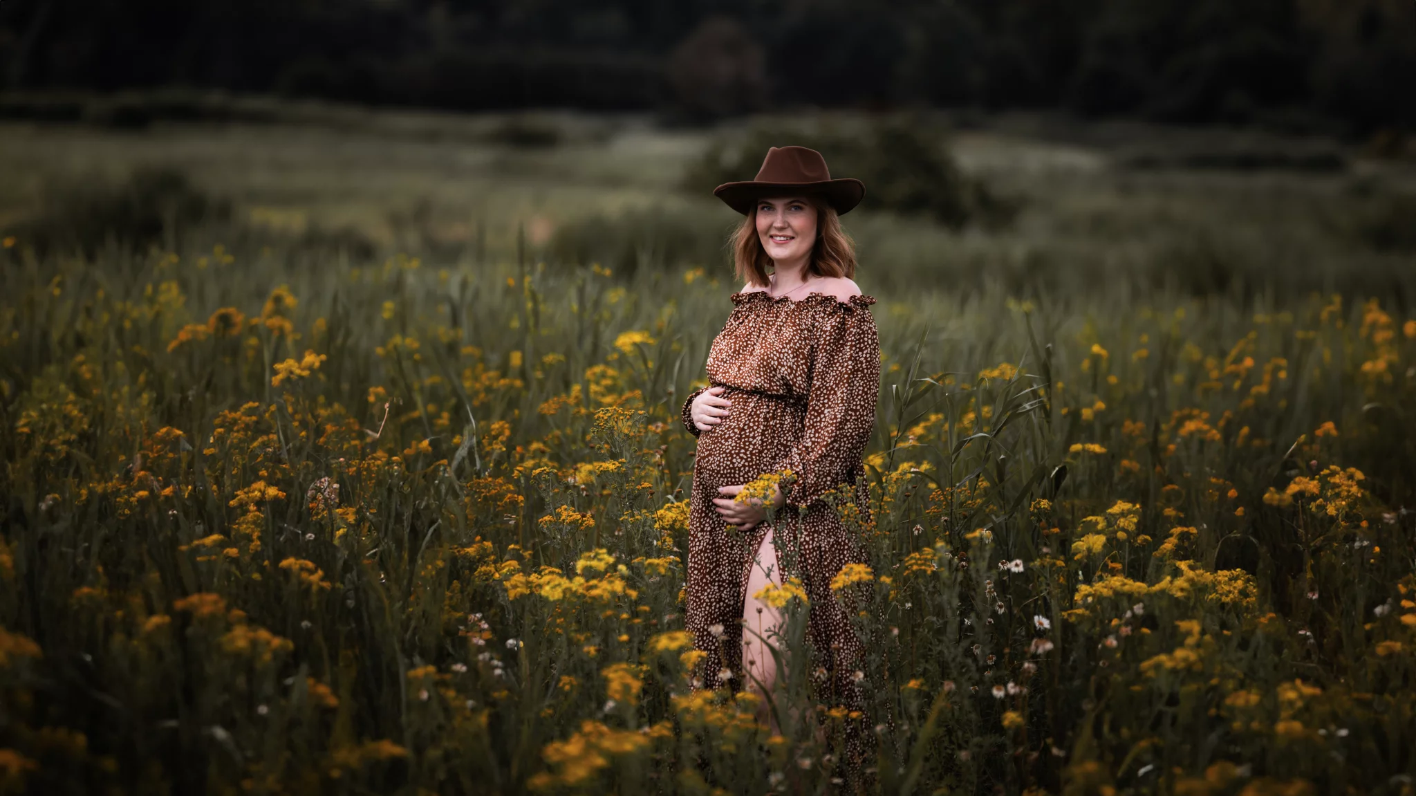 Een zwangere vrouw staat in een veld vol gele bloemen, gekleed in een bruine jurk met een patroon en een brede hoed. Ze glimlacht en houdt haar buik liefdevol vast. De achtergrond is een weids landschap met weelderige vegetatie en donkere bosrand, wat een rustige en natuurlijke sfeer creëert.