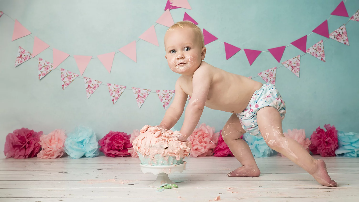 Een baby in een kleurrijke luier staat op een houten vloer en leunt tegen een grote, rommelige taart met roze glazuur. De baby heeft glazuur op zijn gezicht en handen en kijkt met een nieuwsgierige blik naar de camera. Op de achtergrond hangen roze en bloemen vlaggetjes, en op de vloer liggen gekleurde papieren bloemen, wat een feestelijke sfeer creëert. De lichtblauwe achtergrond contrasteert mooi met de licht roze en donker roze decoraties.