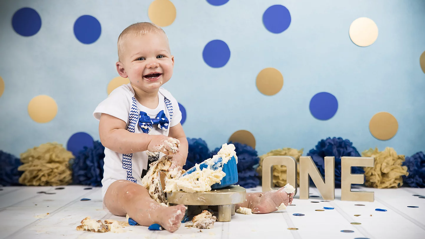 Een vrolijke baby zit op een houten vloer en is bedekt met taartglazuur. De baby draagt een wit shirt met een blauwe strik en bretels en lacht breed naar de camera. Voor de baby staat een deels verpletterde taart, en op de achtergrond zijn blauwe en gouden stippen op een lichtblauwe muur te zien. Achter de baby liggen gekleurde papieren bloemen in blauw en goud, en naast de baby staan grote letters die het woord "ONE" vormen, wat de viering van de eerste verjaardag aangeeft. De sfeer is feestelijk en speels.