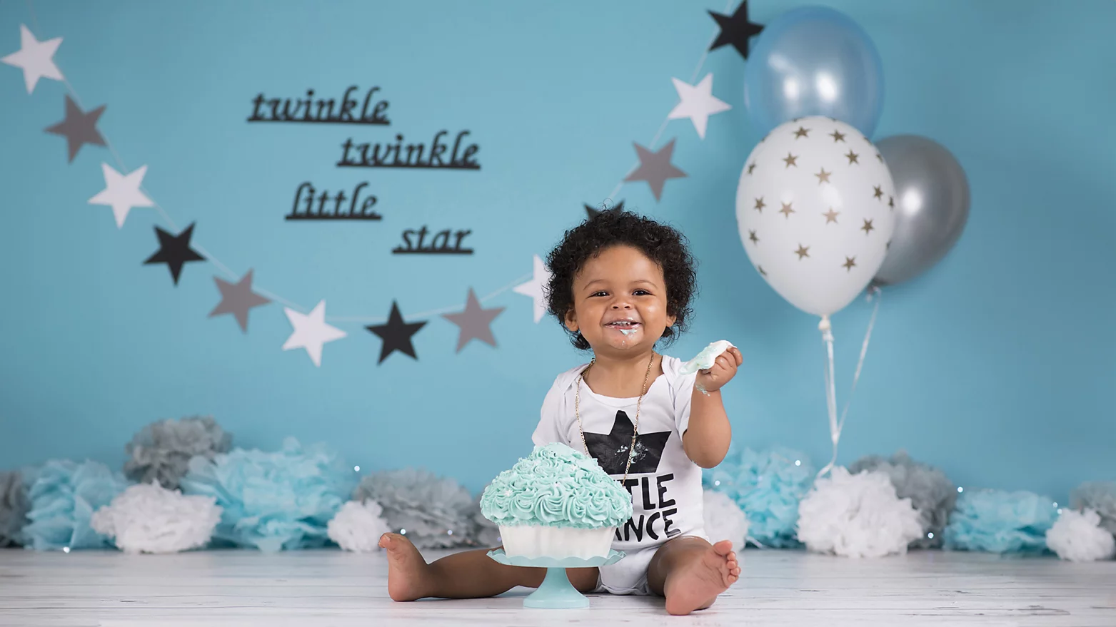 Een vrolijke baby zit op een houten vloer, omringd door feestelijke decoraties. De baby draagt een wit rompertje met de tekst "Little Prince" en heeft een ketting om zijn nek. Voor hem staat een grote cupcake met blauw glazuur, waarvan hij geniet. De achtergrond is lichtblauw met zilveren en blauwe sterren en ballonnen, en de tekst "twinkle twinkle little star" hangt aan de muur. De decoraties omvatten ook papieren pompons in blauw, wit en grijs, wat een feestelijke sfeer creëert.