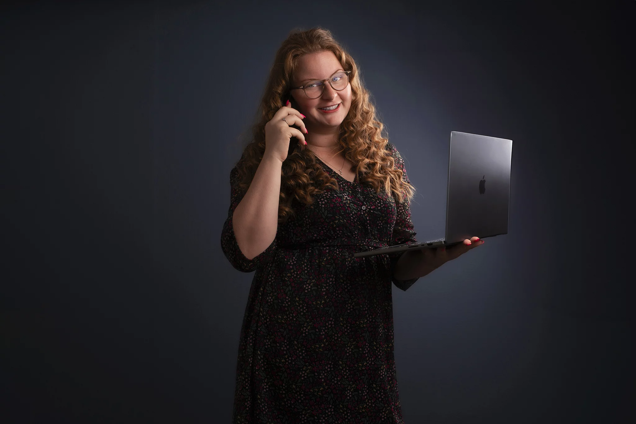 Een vrouw met lang, krullend rood haar en een bril houdt een laptop in haar ene hand en een telefoon tegen haar oor in de andere hand. Ze draagt een donkergekleurd, gebloemd jurkje en glimlacht naar de camera. De achtergrond is donker, wat de aandacht op de vrouw en haar apparatuur vestigt.