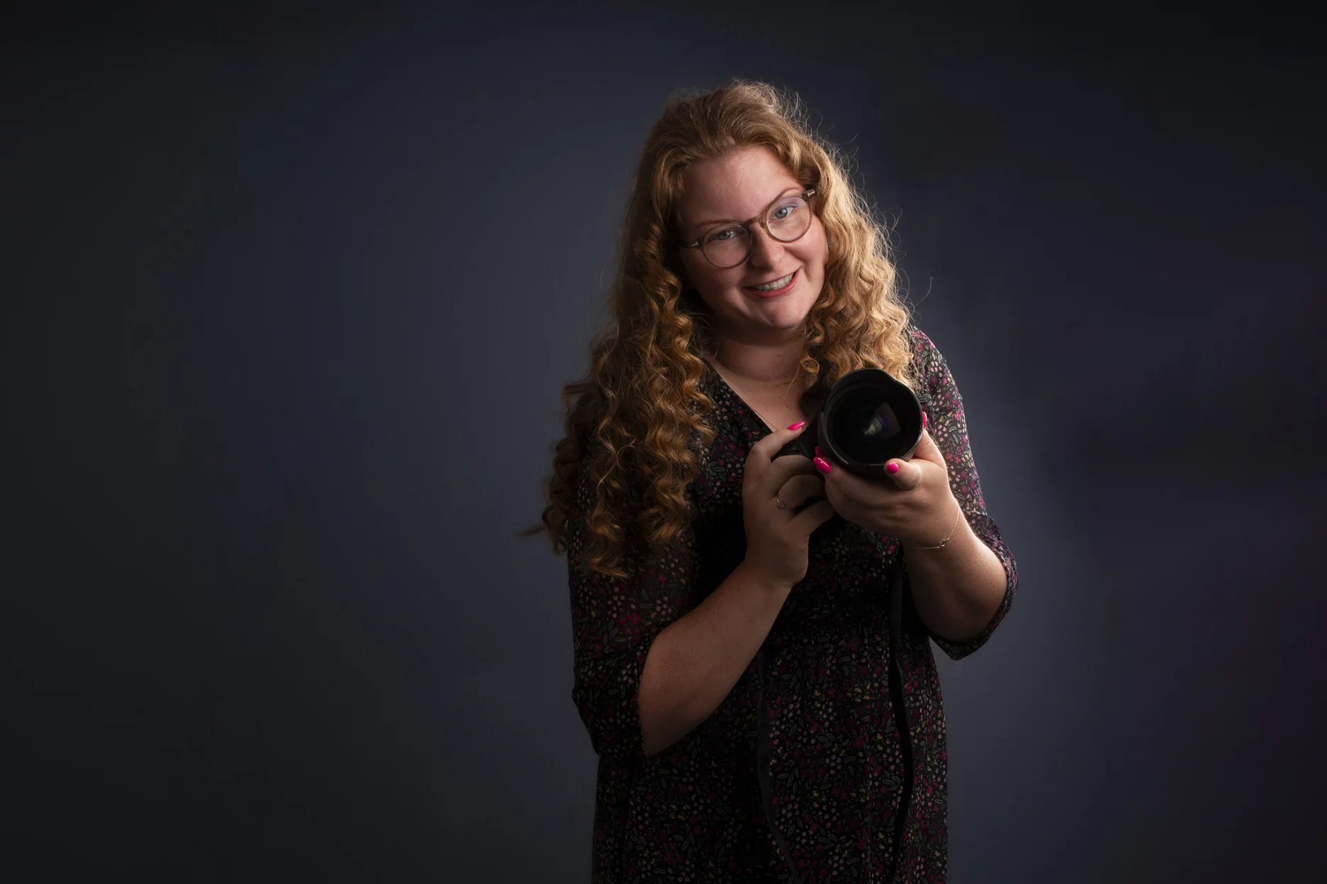 en vrouw met lang, krullend rood haar en een bril houdt een professionele camera vast en kijkt glimlachend naar de camera. Ze draagt een donkergekleurd, gebloemd jurkje. De achtergrond is donker, wat de nadruk legt op de vrouw en haar camera.