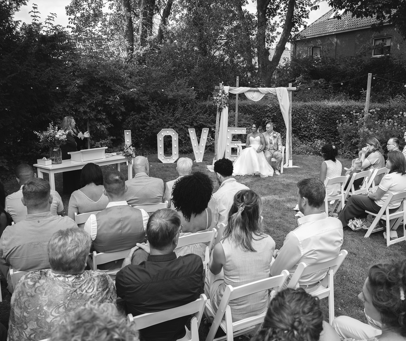 Zwart wit foto van een bruidspaar dit zit onder een boog tijdens de ceremonie in de tuin.